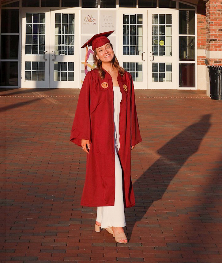 Emma Leonard '25 stands on red brick wearing her cap and gown on graduate day