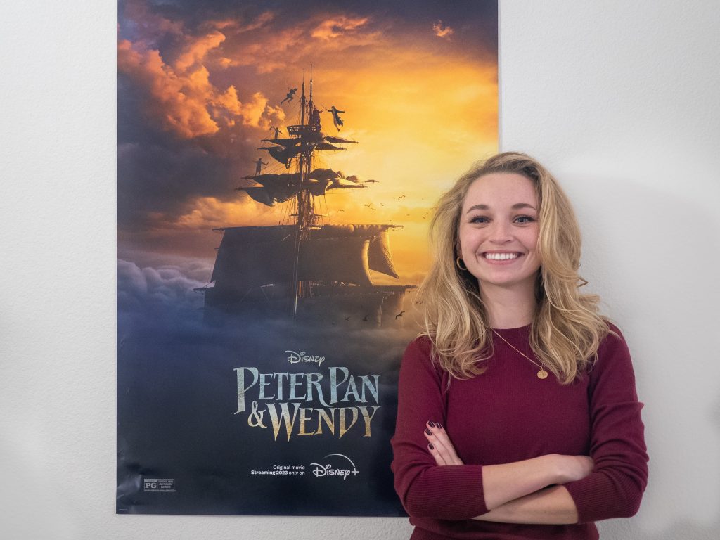 Lilian Engel, smiling with her arms crossed next to a film poster for the 2023 film Peter Pan & Wendy. She served as an apprentice editor for this film.