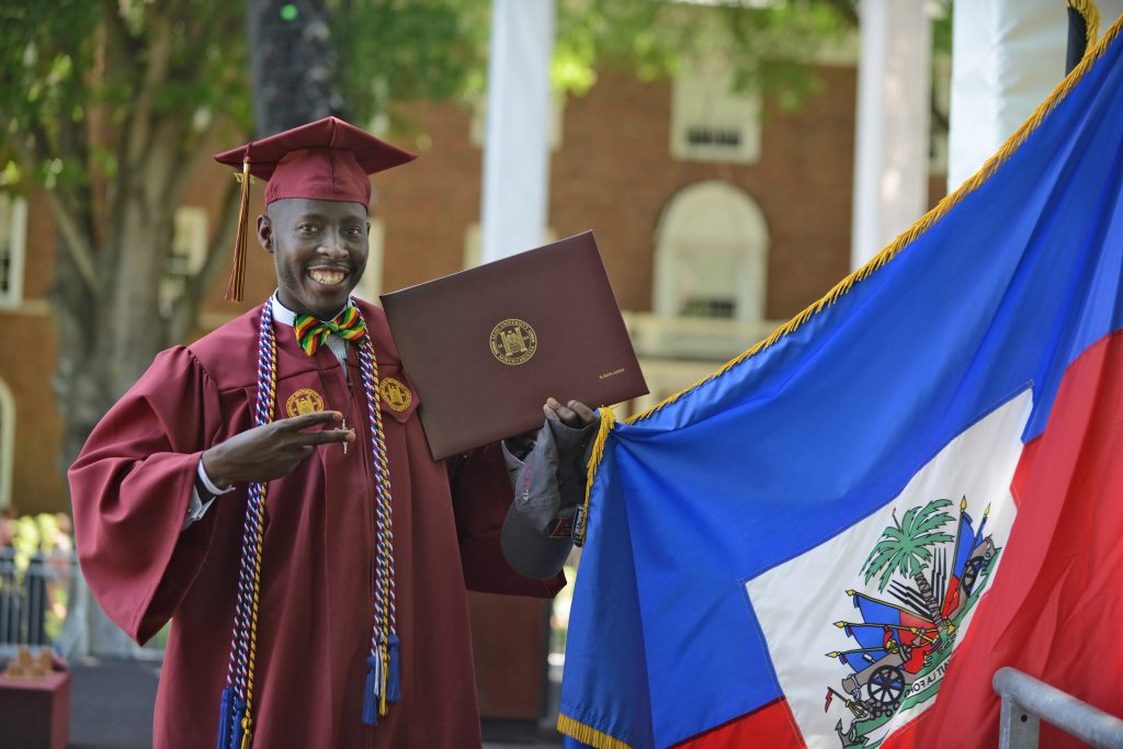 Ellison Adrien, an Elon graduate who majored in biology and is dressed in his Elon graduation garb, points to his diploma.