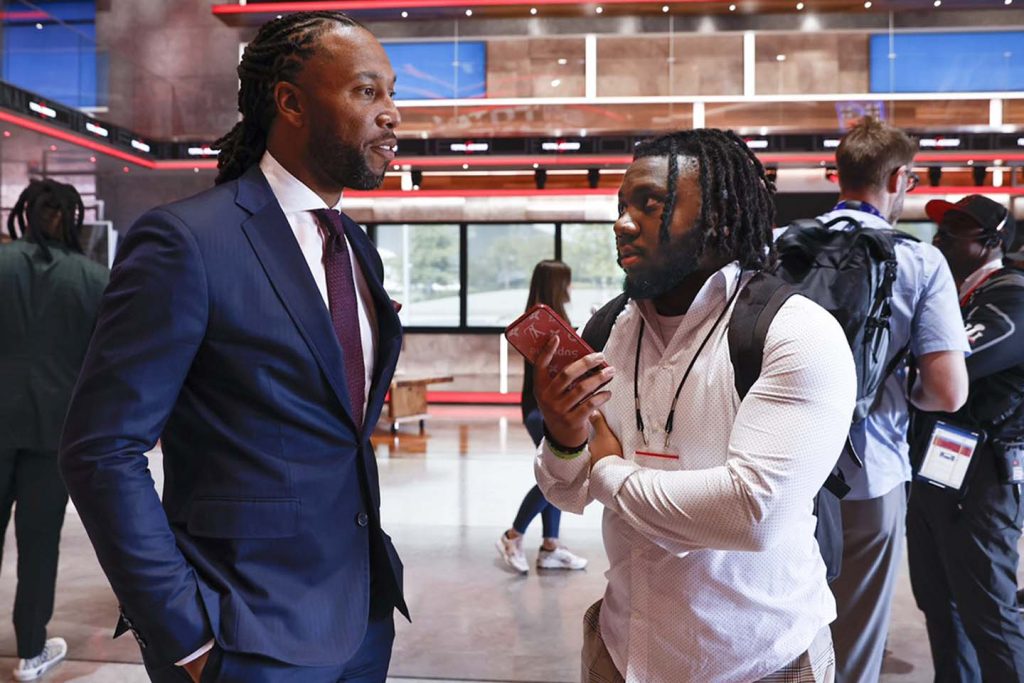 Emmanuel Morgan, an Elon journalism major, interviews NFL legend Larry Fitzgerald during a busy event with people bustling around them.