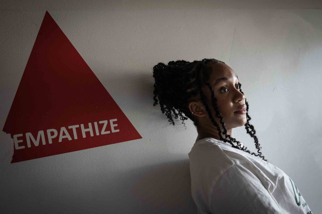 Kiara Hunter is wearing a white T-shirt and standing against a white wall with a red triangle that says empathize.