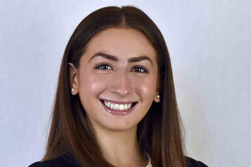 A headshot of Ava Rosen. She has long brown hair and is wearing pearl earrings.