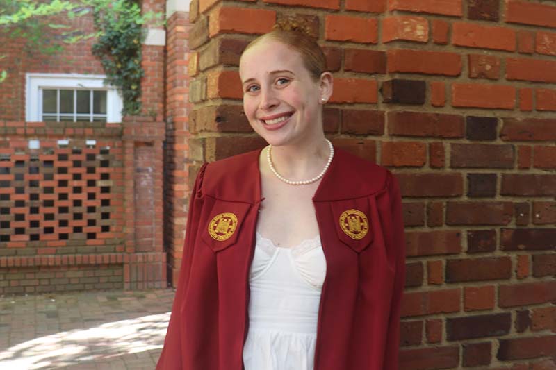 Chloe Stuart stands in front of a brick wall at Elon. She is wearing a maroon graduation gown, a white dress and pearls.