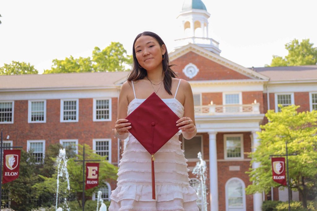 Chloe Yoon is wearing a white dress and holding her Elon graduation cap in front of Alamance building.