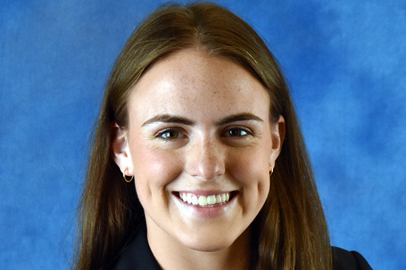 A headshot of Emily Alps. She has long brown hair and is wearing hoop earrings. She is smiling and has dimples.