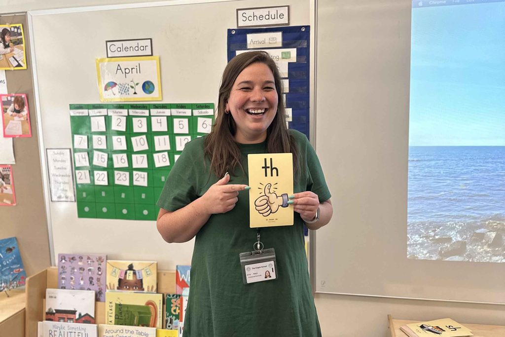 Jenna Mason stands in an elementary school classroom holding a sign that has a drawing of the a thumb and th.
