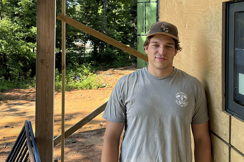 Kade Lervolino is standing next to a tiny house under construction. He is wearing a gray t-shirt and a cap.