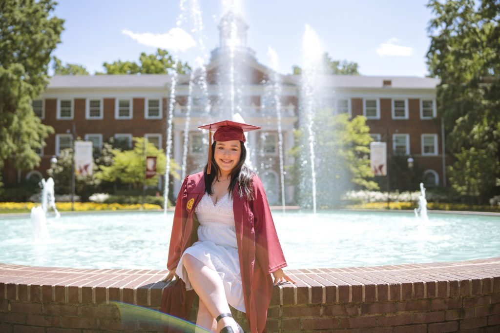 Dressed in a white dress and maroon cap and gown, Maddy Star sits on the brick ledge around Fonville Fountain with Alamance building in the background.