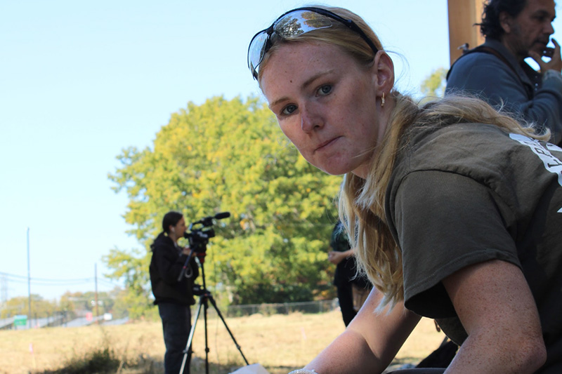 MaryKate Hart is working on a tiny house. She is wearing a t-shirt and sunglasses on the top of her head. There is someone with a video camera in the background.