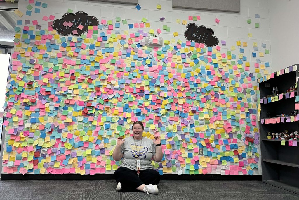 Samantha Childers sits in front of a Gratitude Wall covered in colorful Post-It Notes.