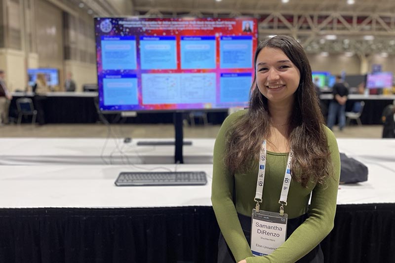 Samantha DiRenzo stands in front of a large computer monitor. She is wearing a nametag around her neck.