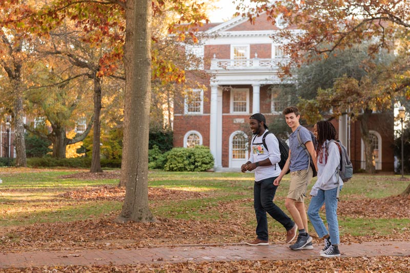 Three students walking along a brick walkway in front of a brick building on Elon's campus.
