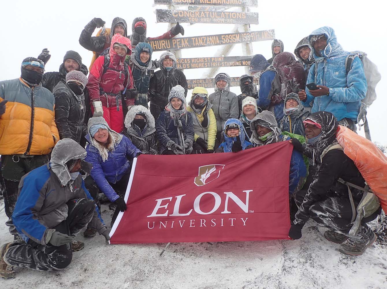 Students participate in one of Elon University's best study abroad programs in Tanzania. They stand at the top of Mount Kilimanjaro holding an Elon University banner, bundled up and surrounded by snow.