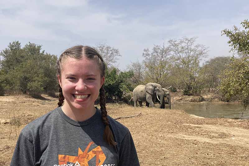 Elon student standing on dirt in Ghana with elephants drinking from a pond in the background
