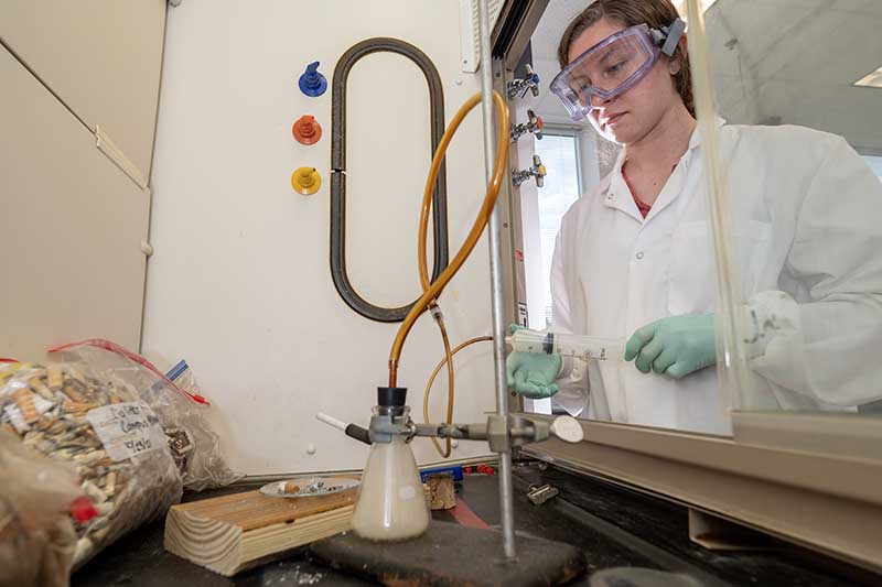 A student wearing goggles and a lab coat operates a smoking device she created in a laboratory. Students are engaged through experiential learning programs like undergraduate research..