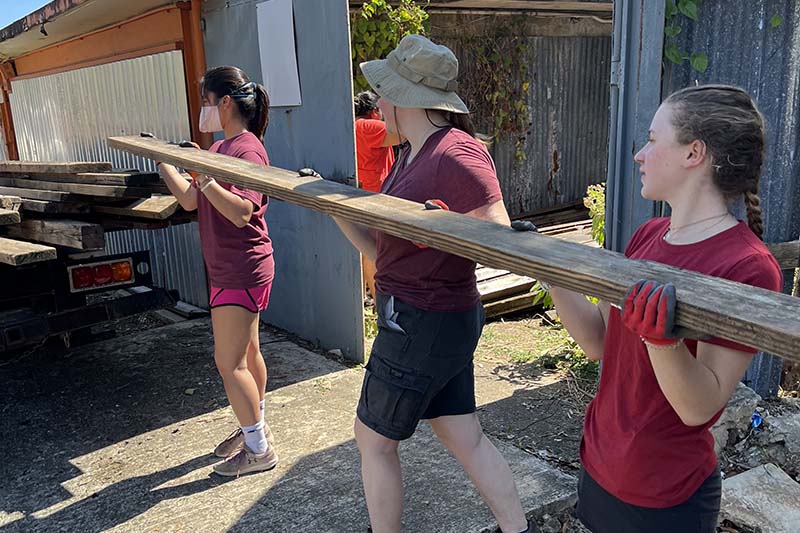 Three students dressed in maroon carry a board as part of a cleanup effort in Puerto Rico following Hurricane Fiona. Students are engaged through experiential learning programs like service..