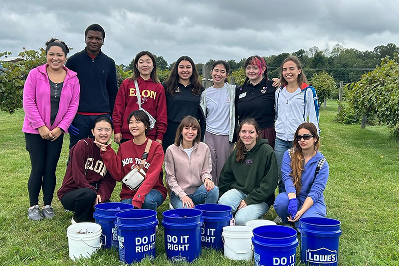 Student volunteers gather for a photo in a blueberry field and have large blue buckets in front of them.