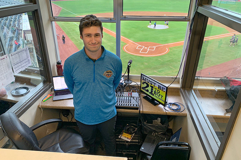Javik Blake stands in the announcer's booth of the Biloxi Shuckers with home plate behind him. Javiks firsthand experiences and internship opportunities at Elon led to his full-time job announcing for the Double A affiliate of the Milwaukee Brewers.