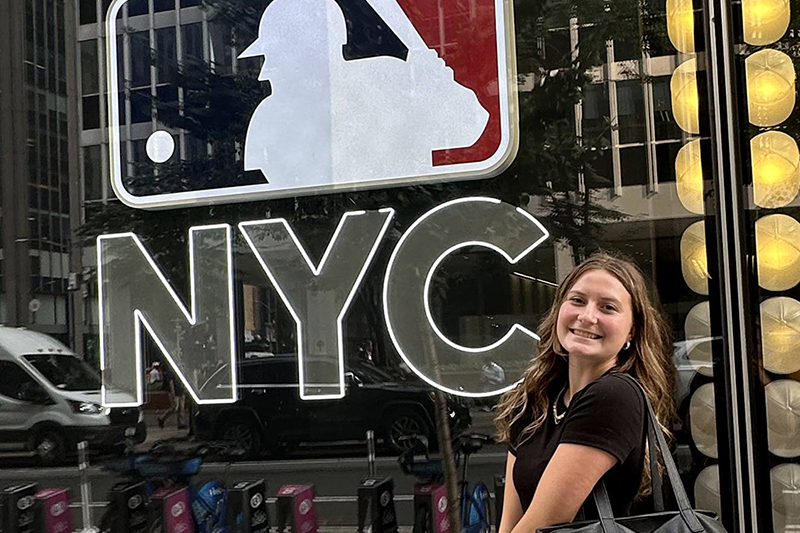 Emilia Cappellett stands in front of a window that has the MLB logo and NYC on it.
