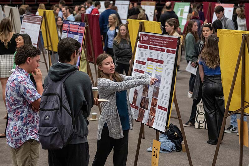 Elon students explain their undergraduate research using posters on easels.