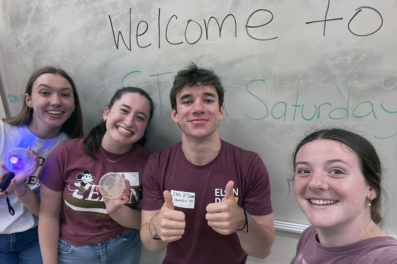 Chris is in a selfie photo with three fellow students and they are standing in front of a hand written STEM Saturday sign.