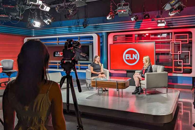 Ginna Claire Mason '13 is interviewed by Anjolina Fantaroni on the Elon News Network set. There are lights hanging from the ceiling. A person is operating the video camera.