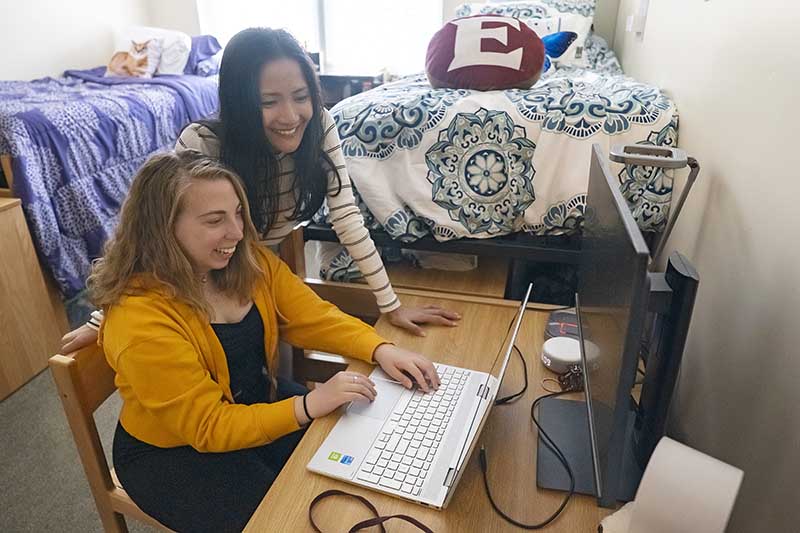 Two roommates in a Colonnades Neighborhood double room stare at a laptop together. Their beds, one with a purple comforter and the other with an Elon pillow on it, stand in the background.