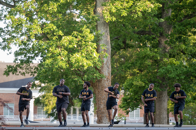 Six men black Alpha Phi Alpha Fraternity t-shirts practice for a step show in the Lambert Academic Village