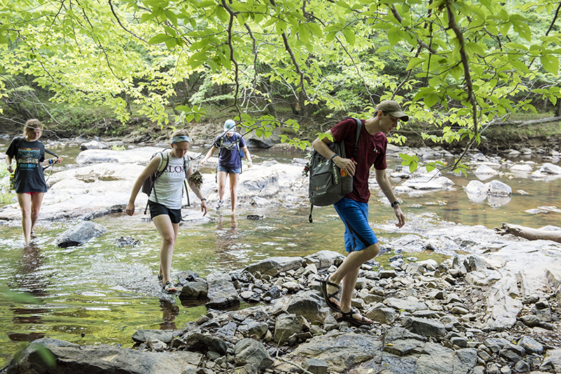 Incoming first-year students attend session two of Adventures in Leadership. A group of students wearing backpacks hikes across rocks in the Eno River.
