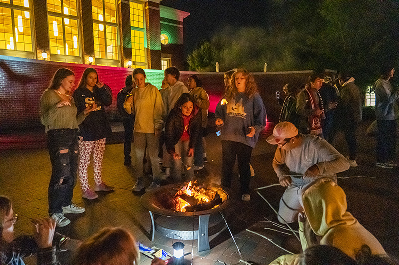 Students gather around an outdoor fire pit and toast marshmallows on sticks.