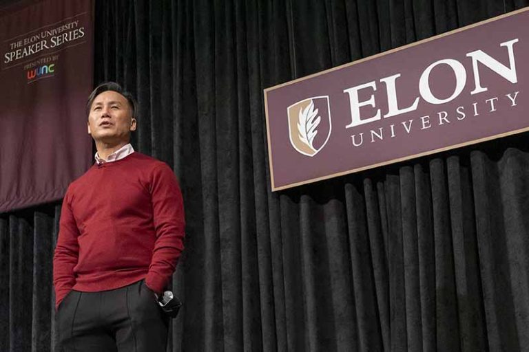 BD Wong, dressed in a red sweater and black pants, stands on an Elon stage speaking with a black curtain with an Elon University sign and Speaker Series sign on it in the background.