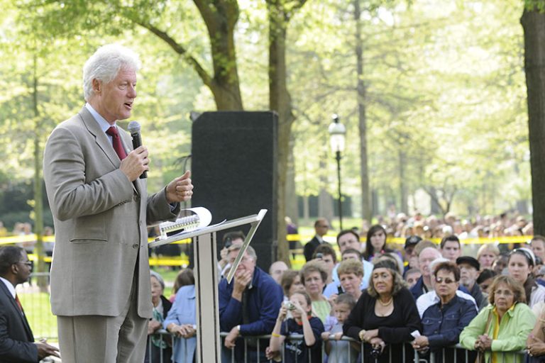 Former President Bill Clinton speaks to a crowd in front of Alamance Building. He is wearing a light suit and a red tie and is holding microphone.