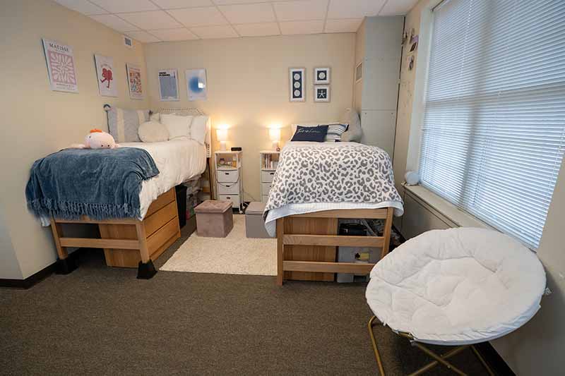 A double room in the Colonnades neighborhood. There are two beds with dressers underneath, identical storage boxes and night stands and comfortable white chair.
