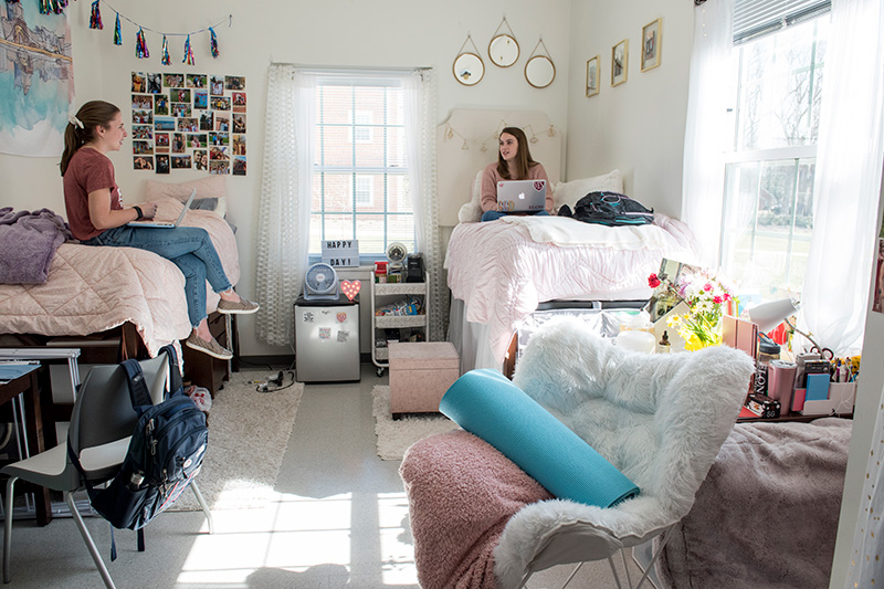 Two roommates in East Residence Hall sit on their beds and chat. The room is filled with typical college room items such as a mini refrigerator, desks, a fuzzy chair and walls covered in photos.