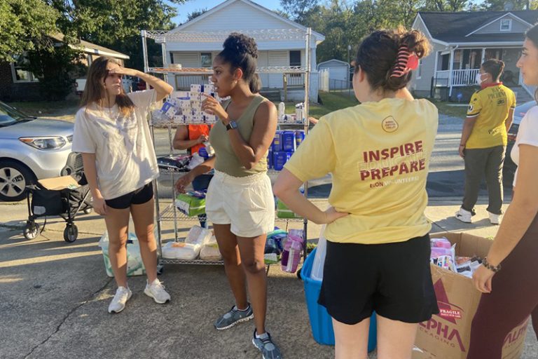 Students in Elon Volunteers! chat at an event. They are standing outside with a shelf and a cardboard box that have items in them.