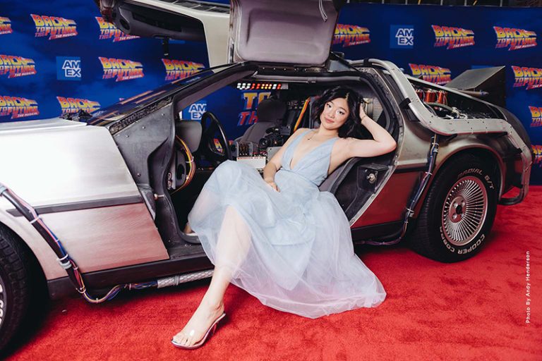 Hannah Kevitt, who is wearing a flowing light blue chiffon dress, sits in a car on a red carpet with Back to the Future backdrop.
