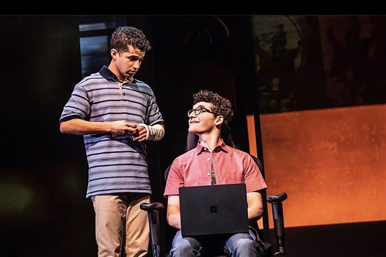 Jared Goldsmith playing the role of Jared Kleinman in the Broadway production of "Dear Evan Hansen." Jared is sitting on stage with a laptop looking at Evan Hansen who is wearing a striped blue shirt and has a cast on his arm.
