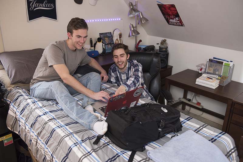 Two roommates are looking at a red laptop and smiling. One of them is on the bed. The other is in a chair beside the bed.