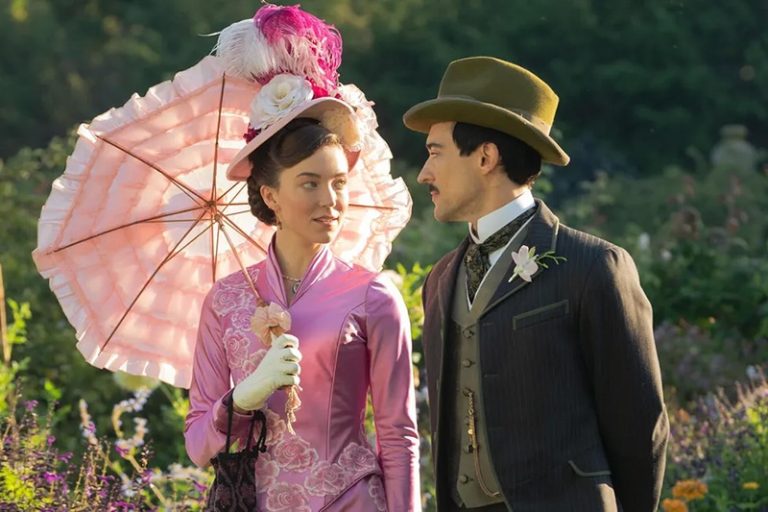 Nicole Brydon Bloom playing the role of socialite Caroline Stuyvesant in the HBO series "Gilded Age." She is wearing a pink dress from that time period, a hat and carrying a pink parasol. She is walking with Blake Ritson, a British actor who portrays Oscar van Rhijn. He is wearing a suit and hat.