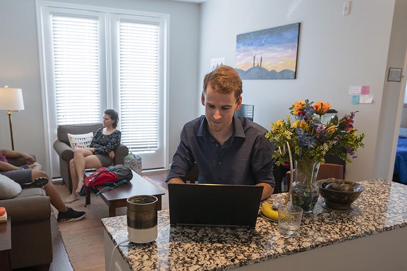A resident of Park Place works on his laptop at the counter in his apartment. There is a vase of flowers, a bowl of fruit and a banana on the counter. Another roommate chats with a visitor on the couch in the background.