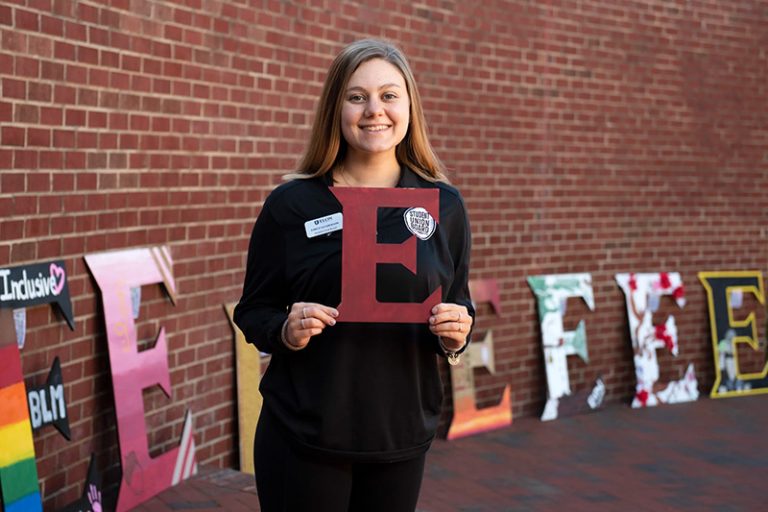 A member of the Student Union Board holds a maroon E. Behind her, there are large monogram Es decorated in a variety of ways. They are leaning against a brick wall.