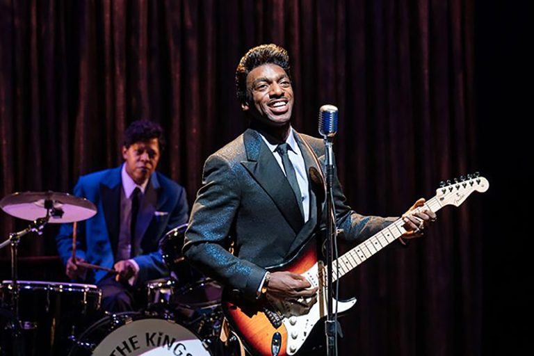 Daniel J. Watts as Ike Turner playing a guitar in the "Tina: The Tina Turner Musical" and singing into a microphone. Another actor in a blue jacket plays the drums behind him.