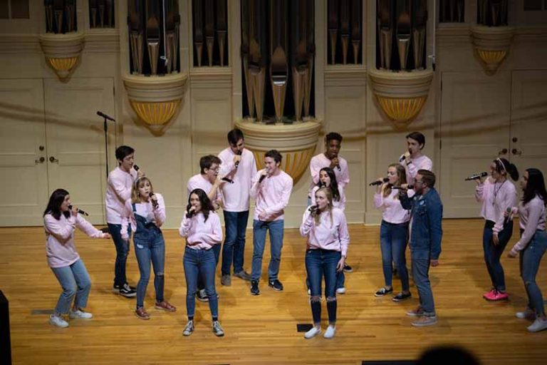 An a capella group dressed jeans and pink shirts perform on the Whitley Auditorium stage.te