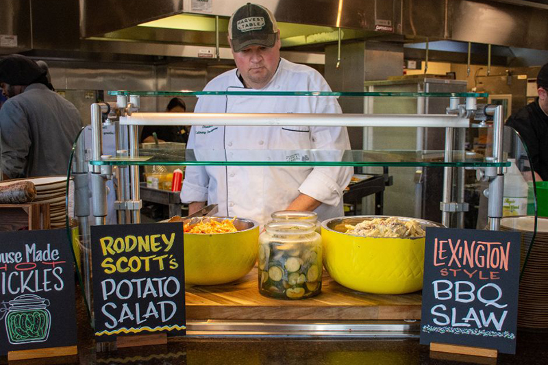 A chef in chef's coat stands at a station in Clohan Hall that serves Lexington style BBQ slaw, potato salad and house made pickles