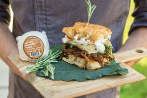 A chef holds a cutting board that has Elon's Bison Bliss, which is a sandwich with locally sourced bison, smoked goat cheese, kale, caramelized onions and rosemary on scratch made focaccia. There is also a packed smokey mountain round cheese and rosemary sprigs on the board.