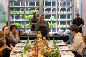 A chef prepares a farm table dinner for students with the herb garden in McEwen Dining Hall behind them.