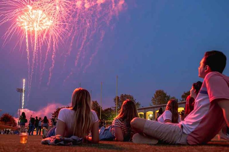 Students are sitting on the ground and looking up at the sky at purple and pink fireworks.