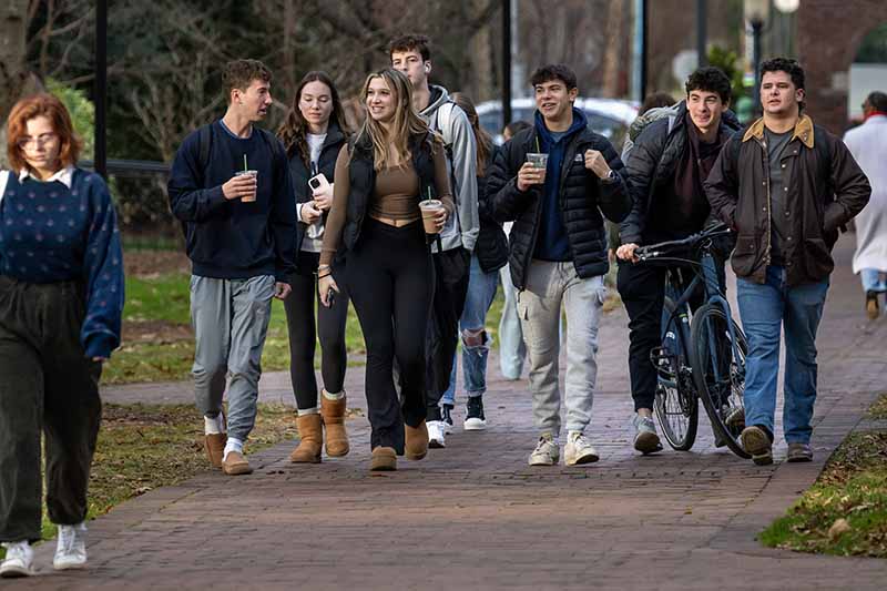A group of students are walking together on campus. Several are holding iced coffee. One has a bike.