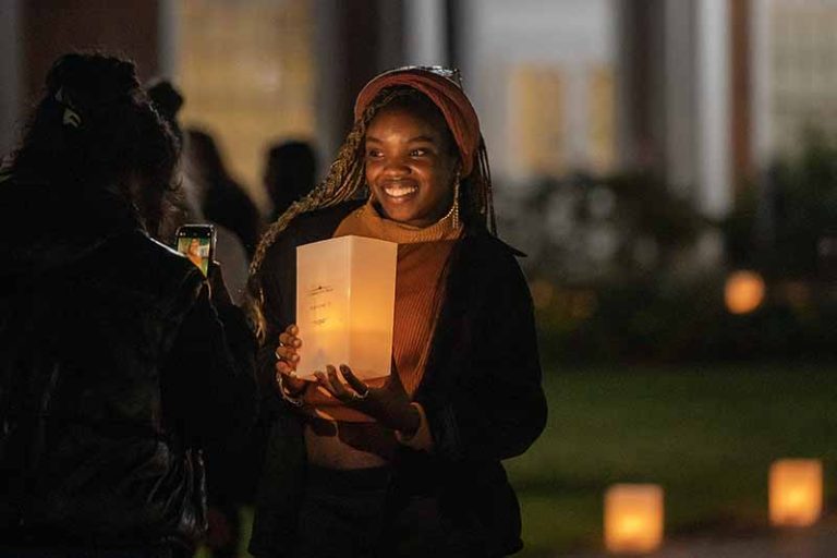A student with braids holds up a lit luminary while another student takes her photo with a mobile phone.