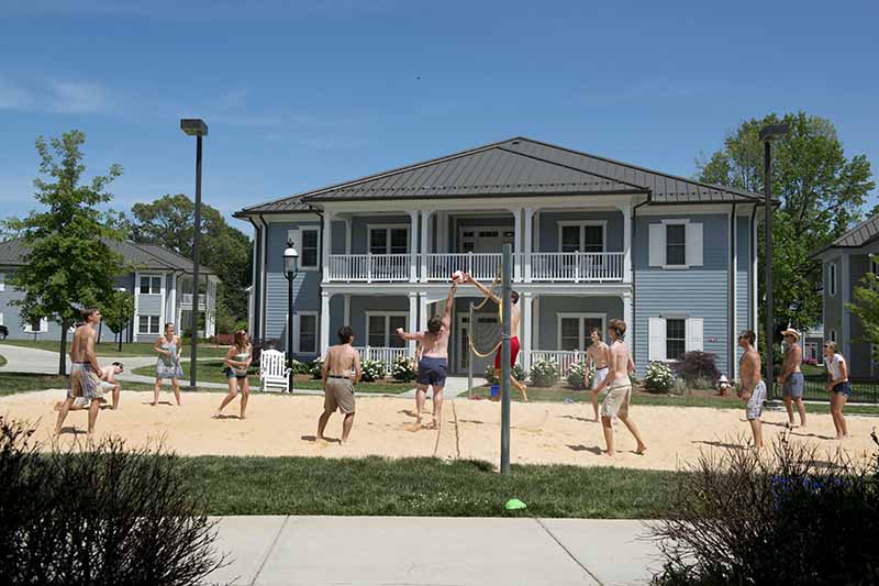 Residents of Station at Mill Point Neighborhood play volleyball in the sand. The guys are all shirtless. One guy is at the net trying to tap the ball over while his opponent blocks it.